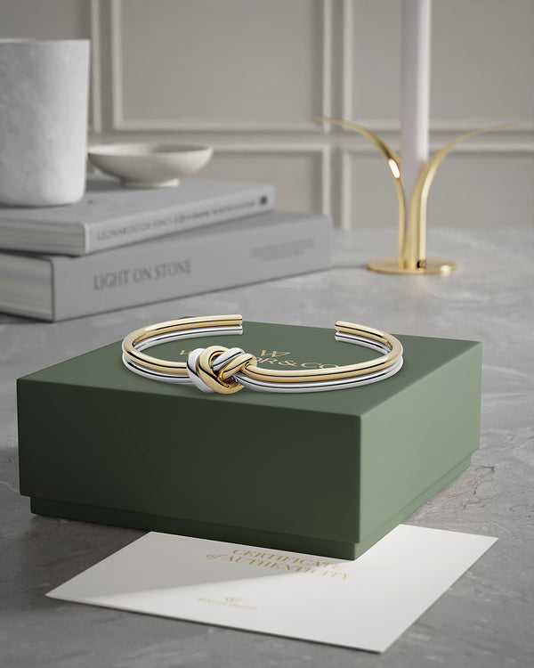 A polished stainless steel bangle in 14k gold from Waldor & Co. One size. The model is Dual Knot Bangle.